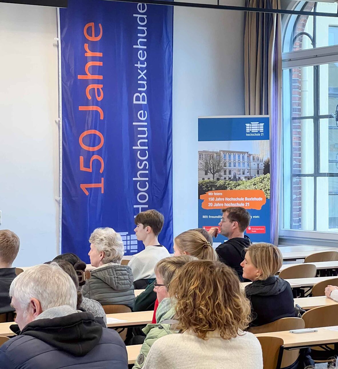In der Aula, in der eine Fahne mit dem Schriftzug 150 Jahre Hochschule Buxtehude hängt, hören Besucherinnen und Besucher einem Vortrag zu