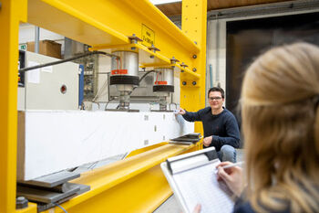 Unscharf im Vordergrund eine Person schreibt auf ein Klemmbrett. Im Hintergrund ein Mann neben einer Maschine