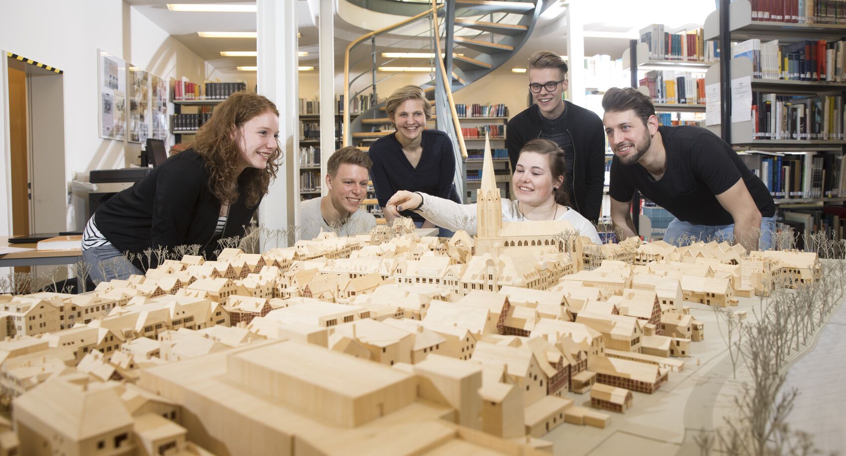 Gruppe junger Menschen betrachtet ein Miniatur-Stadtmodell aus Holz, das in der Bibliothek steht