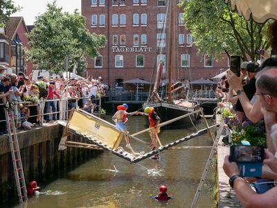 Zwei Studierende stehen auf einer Holzbrücke, die gerade einstürzt. Viele Schaulustige stehen am Geländer.