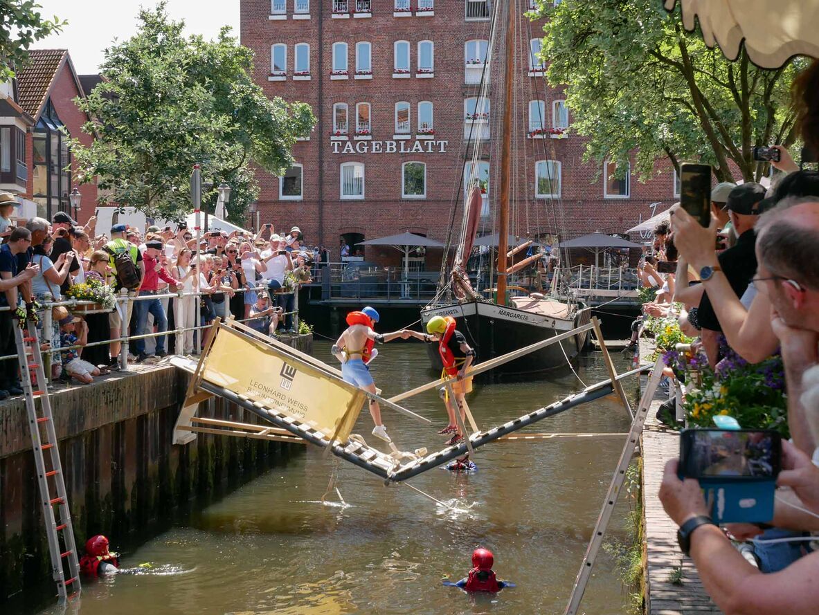 Zwei Studierende stehen auf einer Holzbrücke, die gerade einstürzt. Viele Schaulustige stehen am Geländer.