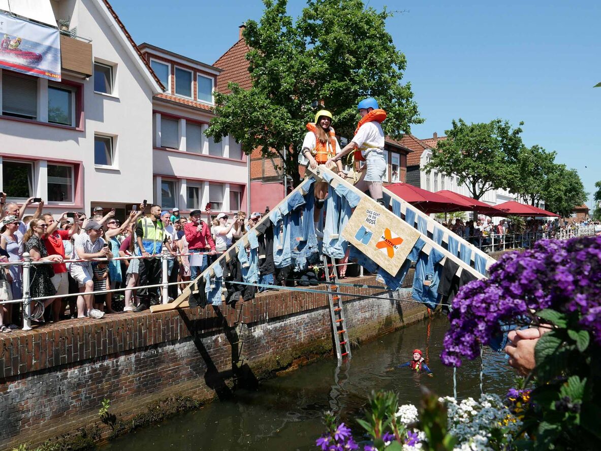 Zwei Studierende stehen auf einer Brücke, aus Jeans und Holzbohlen gefertigt, über dem Fleth und geben sich die Hand, während viele Zuschauer zusehen.